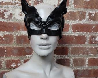 May include: A black leather mask with pointed ears and a sculpted face design. The mask covers the eyes and nose, with a detailed, textured appearance. The mask is displayed on a mannequin against a brick wall.