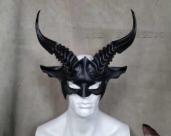 Pode incluir: Uma máscara preta com chifres com um design detalhado, com chifres curvos e uma cobertura facial. A máscara é exibida em uma cabeça de manequim branca, mostrando seu artesanato intrincado. O design da máscara sugere um tema de fantasia ou teatral.