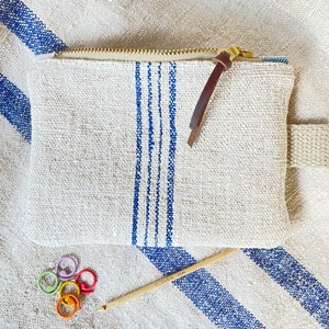 May include: A white linen zippered pouch with blue stripes and a brown leather zipper pull. The pouch is lying on a white and blue striped fabric with a wooden knitting needle and colorful stitch markers.