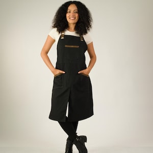 May include: A black apron with a white t-shirt underneath. The apron features a square chest pocket, two large hip pockets, and brown leather-like straps with metallic buckles. The design includes a central vertical slit at the bottom hem.