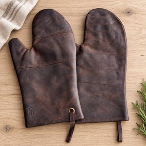 May include: A pair of dark brown leather oven mitts featuring horizontal stitching across the top section, a smooth lower palm area, and a small embossed leaf logo. Each mitt includes a brass-toned metal grommet with a leather hanging loop.