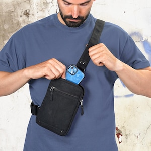May include: Black canvas crossbody bag with a zippered main compartment and an adjustable strap. A blue smartphone is visible inside the bag's pocket. The bag is worn over the shoulder.