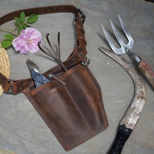 May include: A brown leather gardening tool belt with multiple pockets and a metal ring. The belt is worn over a person's shoulder and has a variety of gardening tools attached to it, including a pair of pruning shears, a hand rake, and a trowel.
