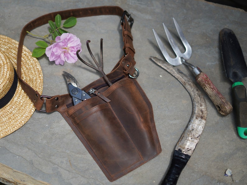 May include: A brown leather gardening tool belt with multiple pockets and a metal ring. The belt is worn over a person's shoulder and has a variety of gardening tools attached to it, including a pair of pruning shears, a hand rake, and a trowel.