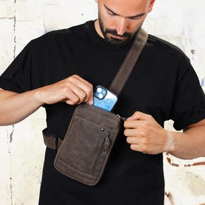 May include: A brown leather crossbody bag with a front zipper pocket and adjustable strap. The bag is worn across the body and has a phone in the main compartment. The bag is made of a distressed leather material.