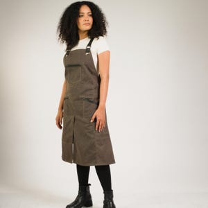 May include: A dark gray apron featuring a central chest pocket and two large side pockets, all accented with black leather trim. The apron has adjustable black leather shoulder straps with metal buckles and a vertical slit at the bottom hem.