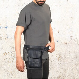 Puede incluir: Ri&ntilde;onera de cuero negro con un bolsillo delantero y un dise&ntilde;o tejido. La ri&ntilde;onera se lleva alrededor de la cintura y tiene un cierre de hebilla. La persona de la imagen lleva una camiseta gris y pantalones oscuros.