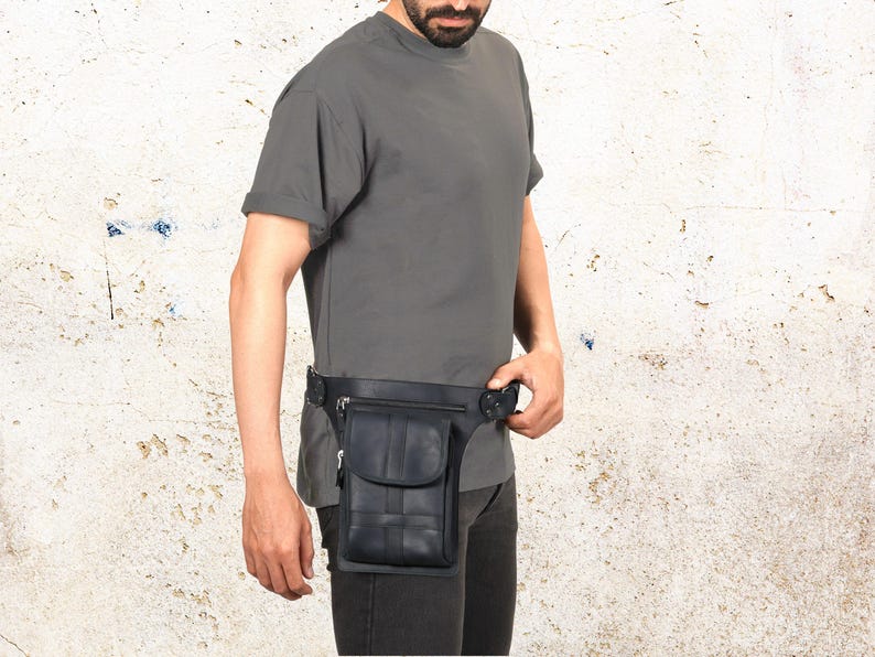 Puede incluir: Ri&ntilde;onera de cuero negro con un bolsillo delantero y un dise&ntilde;o tejido. La ri&ntilde;onera se lleva alrededor de la cintura y tiene un cierre de hebilla. La persona de la imagen lleva una camiseta gris y pantalones oscuros.