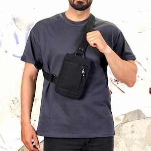 Könnte beinhalten: Schwarze Umhängetasche mit Reißverschluss-Vordertasche und verstellbarem Riemen. Die Tasche wird quer über dem Körper getragen. Die Person trägt ein dunkelgraues T-Shirt und eine schwarze Hose. Der Hintergrund ist eine abgenutzte weiße Wand.