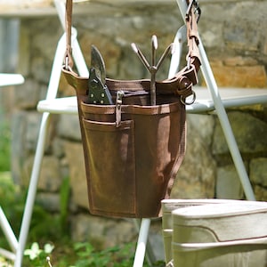 May include: A brown leather gardening tool bag with a shoulder strap. The bag has multiple pockets and is hanging on a white metal chair. The bag contains gardening tools, including a pair of pruning shears and a hand rake.