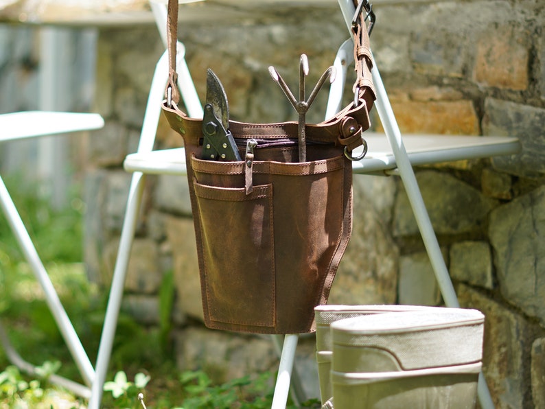May include: A brown leather gardening tool bag with a shoulder strap. The bag has multiple pockets and is hanging on a white metal chair. The bag contains gardening tools, including a pair of pruning shears and a hand rake.