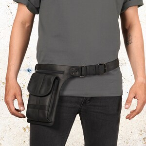 Puede incluir: Cintur&oacute;n de cuero negro con bolsillo y cierre de hebilla. El cintur&oacute;n tiene un bolsillo rectangular en el lado izquierdo con cierre de solapa. El cintur&oacute;n se usa con pantalones negros y una camisa gris.
