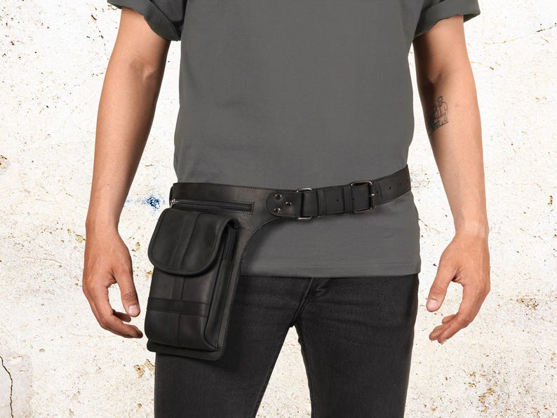 Puede incluir: Cintur&oacute;n de cuero negro con bolsillo y cierre de hebilla. El cintur&oacute;n tiene un bolsillo rectangular en el lado izquierdo con cierre de solapa. El cintur&oacute;n se usa con pantalones negros y una camisa gris.