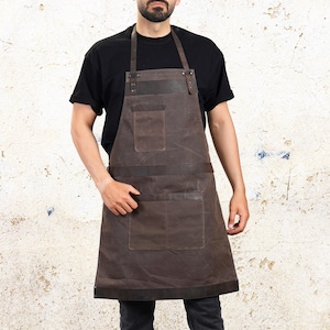 May include: A brown leather apron with two large pockets and leather straps. The apron is worn by a person wearing a black t-shirt. The apron is designed for use in the kitchen or workshop.