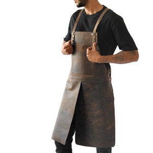 May include: A brown leather apron with adjustable straps and a front pocket. The apron is designed for full coverage and features a split at the bottom for ease of movement. The leather has a distressed finish.