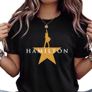 Może przedstawiać: Czarny t-shirt z napisem "HAMILTON" w kolorze białym, nad złotą grafiką gwiazdy z sylwetką osoby. Koszulka jest z kr&oacute;tkim rękawem i wykonana z miękkiego materiału.