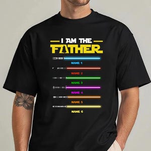 Może przedstawiać: Czarna koszulka z napisem "I AM THE FATHER" w kolorze ż&oacute;łtym i białym. Poniżej sześć mieczy świetlnych z ostrzami w r&oacute;żnych kolorach oraz napisy "NAME 1" do "NAME 6" w kolorze niebieskim, czerwonym, zielonym, fioletowym, pomarańczowym i ż&oacute;łtym.