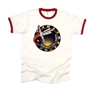 Puede incluir: Camiseta ringer color crema con cuello y ribetes de manga rojos. La camiseta presenta un gr&aacute;fico circular con el texto "PROJECT HAIL MARY" y un dise&ntilde;o de cohete y banderas de varios pa&iacute;ses.