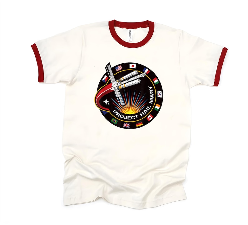 Puede incluir: Camiseta ringer color crema con cuello y ribetes de manga rojos. La camiseta presenta un gr&aacute;fico circular con el texto "PROJECT HAIL MARY" y un dise&ntilde;o de cohete y banderas de varios pa&iacute;ses.