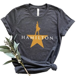 Może przedstawiać: Ciemnoszara koszulka z napisem "HAMILTON" w białych literach nad złotą gwiazdą. Sylwetka osoby stoi na gwieździe. Koszulka jest złożona i zawiązana na dole.