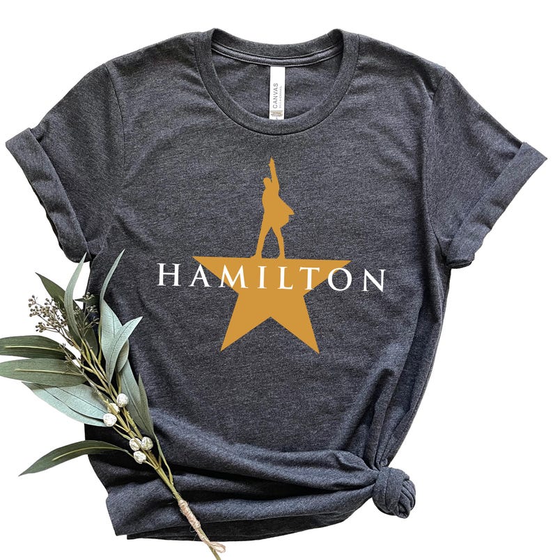 Może przedstawiać: Ciemnoszara koszulka z napisem "HAMILTON" w białych literach nad złotą gwiazdą. Sylwetka osoby stoi na gwieździe. Koszulka jest złożona i zawiązana na dole.