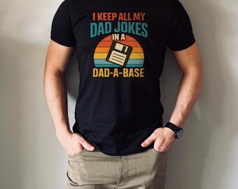 May include: Black t-shirt with the text "I KEEP ALL MY DAD JOKES IN A DAD-A-BASE" in teal, orange, and yellow. The graphic includes a floppy disk design set against a sunset background. The shirt is a classic crew neck.