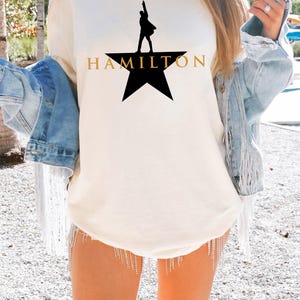 Może przedstawiać: Kremowa koszulka z czarną sylwetką osoby na gwieździe i napisem "HAMILTON" w złotych literach. Koszulka jest częścią kolekcji odzieży.