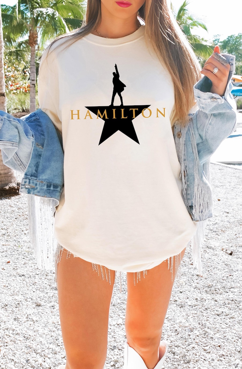 Może przedstawiać: Kremowa koszulka z czarną sylwetką osoby na gwieździe i napisem "HAMILTON" w złotych literach. Koszulka jest częścią kolekcji odzieży.