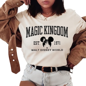 May include: Cream-colored t-shirt with the text "MAGIC KINGDOM" in black block letters. Below the text is a silhouette of Mickey Mouse ears and a castle, with "EST. 1971" and "WALT DISNEY WORLD" printed below. The shirt is paired with denim shorts.