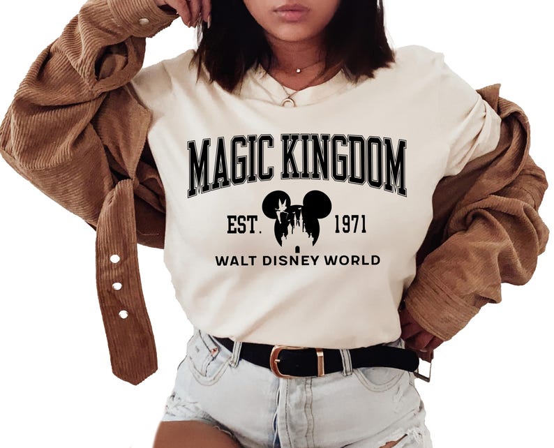 May include: Cream-colored t-shirt with the text "MAGIC KINGDOM" in black block letters. Below the text is a silhouette of Mickey Mouse ears and a castle, with "EST. 1971" and "WALT DISNEY WORLD" printed below. The shirt is paired with denim shorts.