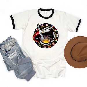Puede incluir: Camiseta blanca con cuello redondo y ribetes negros, con un dise&ntilde;o circular que incluye un cohete estilizado y el texto "PROJECT HAIL MARY", rodeado de banderas. La imagen tambi&eacute;n muestra unos vaqueros azules y un sombrero marr&oacute;n.