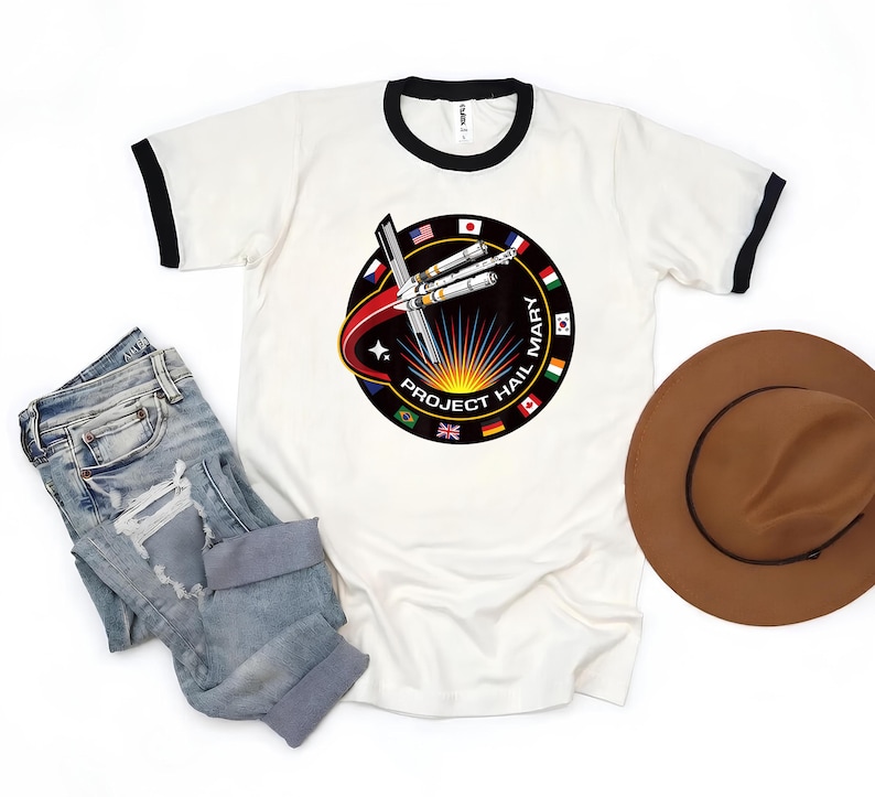 Puede incluir: Camiseta blanca con cuello redondo y ribetes negros, con un dise&ntilde;o circular que incluye un cohete estilizado y el texto "PROJECT HAIL MARY", rodeado de banderas. La imagen tambi&eacute;n muestra unos vaqueros azules y un sombrero marr&oacute;n.