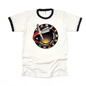 May include: White ringer t-shirt with black trim featuring a circular graphic. The graphic includes a rocket design, the text "PROJECT HAIL MARY", and flags from various countries. The shirt is made of a soft, comfortable material.