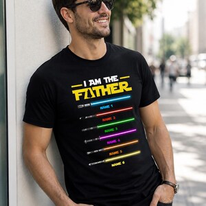 Może przedstawiać: Czarny t-shirt z napisem "I AM THE FATHER" w kolorze ż&oacute;łtym i białym. Poniżej tekstu znajduje się sześć mieczy świetlnych w r&oacute;żnych kolorach, każdy z nazwą. Osoba nosi okulary przeciwsłoneczne.