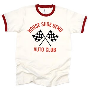 May include: A cream-colored ringer t-shirt with red trim on the collar and sleeves. The front features red text that reads HORSE SHOE BEND AUTO CLUB, positioned above and below two crossed black and white checkered racing flags.