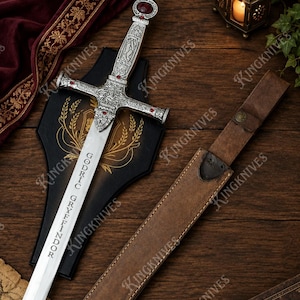 May include: A silver-toned sword with an ornate hilt featuring red gemstone accents and the inscription GODRIC GRYFFINDOR on the blade. The sword is displayed with a black wall plaque and a brown leather scabbard with a metal snap closure.