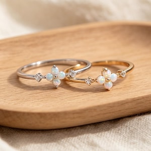 May include: Two thin rings, one in silver-tone and one in gold-tone metal, each featuring a central four-petaled flower design composed of small round opals and flanked by a single tiny clear gemstone on each side of the floral centrepiece.