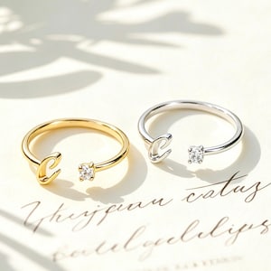 May include: Two open-style rings, one gold-toned and one silver-toned, each featuring a letter C on one end and a small, clear, round-cut gemstone on the other. The rings are placed on a white card with cursive text printed in black ink.