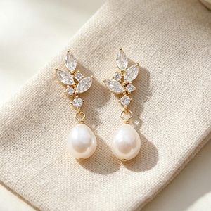 Peut inclure: Paire de boucles d'oreilles pendantes dorées avec un motif en forme de feuille, ornées de cristaux transparents et d'une grosse perle en forme de poire. Les boucles d'oreilles reposent sur un tissu en lin beige.
