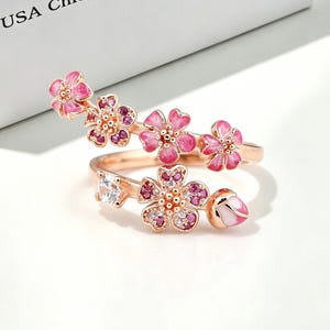 Peut inclure: Une bague en or rose au design ouvert ornée de plusieurs fleurs de cerisier en émail rose, de petits cristaux roses et transparents, et d'un bouton floral. La bague est placée devant une boîte blanche avec le texte USA Chic, RELY RABBIT Unique.