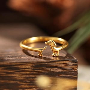 Könnte beinhalten: Ein goldfarbener offener Ring mit einem stilisierten Dackel-Design. Das Ringband bildet den Körper des Hundes, wobei sich Kopf und Vorderpfoten an einem Ende und der Schwanz am anderen Ende befinden, was eine durchgehende Form ergibt.
