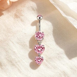May include: A silver-toned navel piercing barbell featuring a top ball and a vertical drop design. The piece is adorned with three faceted, heart-shaped pink stones set in metal prongs, arranged in a descending size order from top to bottom.