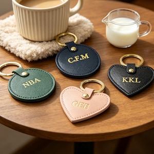 May include: Four leather keychains with gold-tone metal rings and rivets. Two are circular in dark green and navy blue with gold embossed initials NBA and C.F.M. Two are heart-shaped in black and light pink with gold embossed initials KKL and GEE.