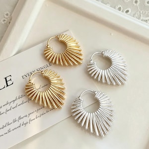 Peut inclure: Deux paires de boucles d'oreilles créoles, une paire dorée et une paire argentée. Chaque boucle présente une base circulaire avec des pointes métalliques texturées en forme d'éventail rayonnant vers l'extérieur, créant un motif en soleil.