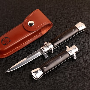 May include: Two switchblade knives with black handles and silver detailing, and a brown leather sheath. The knives are open, showing their blades. The sheath is embossed with the name "Grayson".