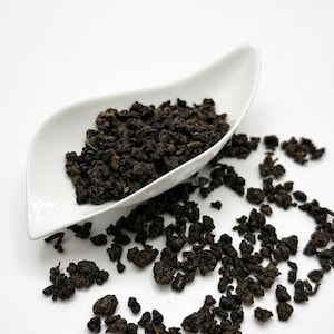 May include: Dark, dried tea leaves in a white, leaf-shaped dish. The tea leaves are a deep brown color and appear to be tightly curled. Some leaves are scattered around the dish on a white surface. The image is well-lit and the tea leaves are in focus.