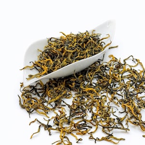May include: Close-up of loose leaf tea with golden and dark green hues, spilling from a white, leaf-shaped dish onto a white surface. The tea leaves have a twisted, wiry texture, suggesting a high-quality product.