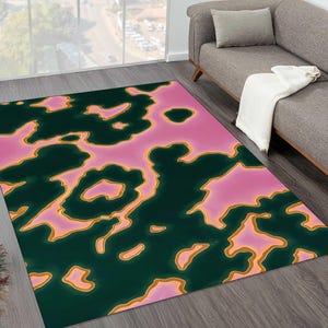 May include: A rectangular area rug with a unique abstract design. The rug features a pattern of dark green shapes outlined in gold against a pink background. The rug is placed on a wooden floor.