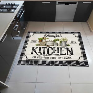 May include: A rectangular kitchen mat with a black and white checkered border. The center features a white wooden plank design with the word "KITCHEN" in large black letters. The mat includes the text "Jennifer's", "Eat Well - Pray Often - Love Always", and the year 2025.