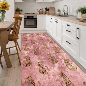 May include: A pink kitchen rug with a leopard print design. The rug features a repeating pattern of leopard spots and figures on a textured pink background. The rug is rectangular and placed in front of a white kitchen cabinet.